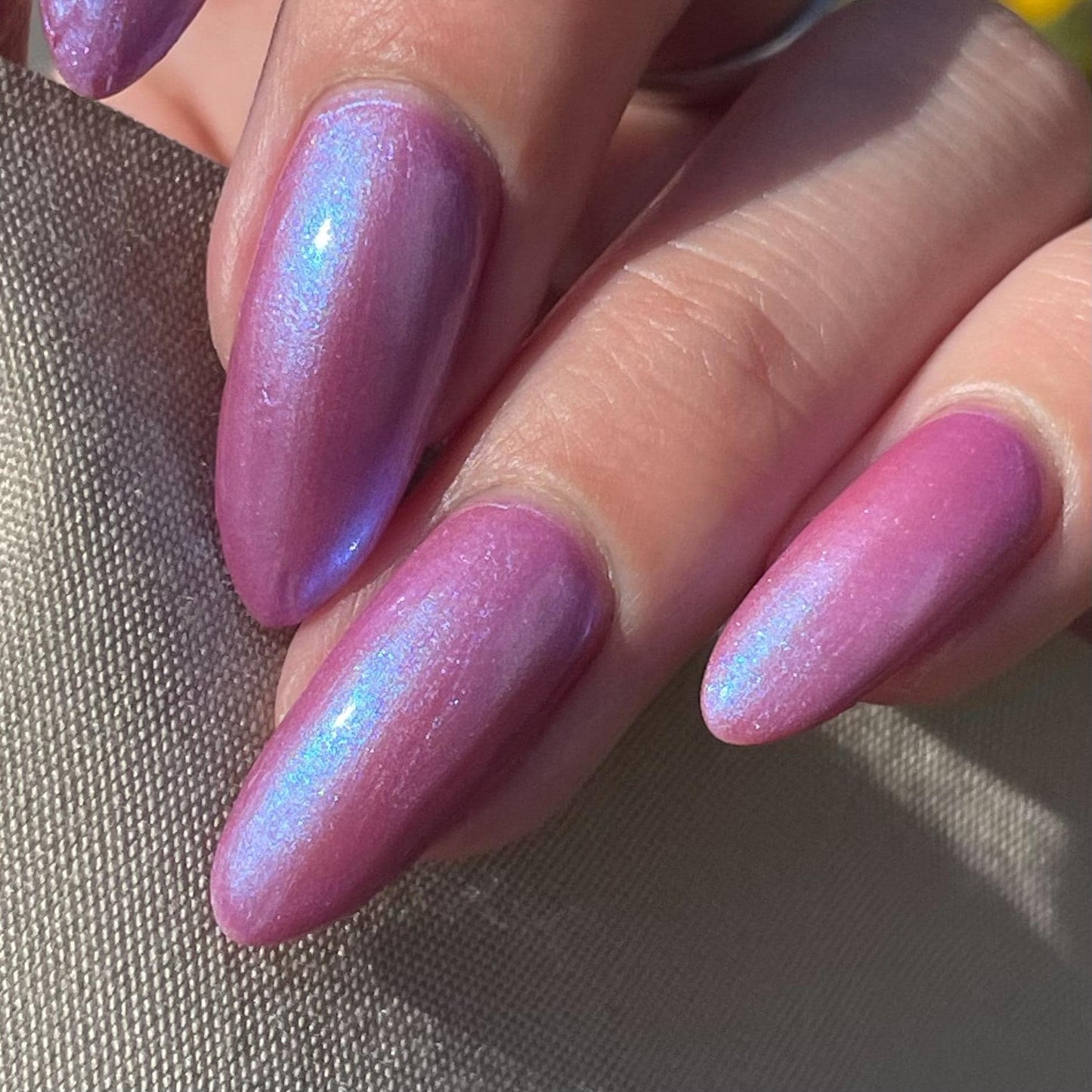 A hand with long nails is painted a purple mauve color with blue shimmer that appears to glow in the sunlight.