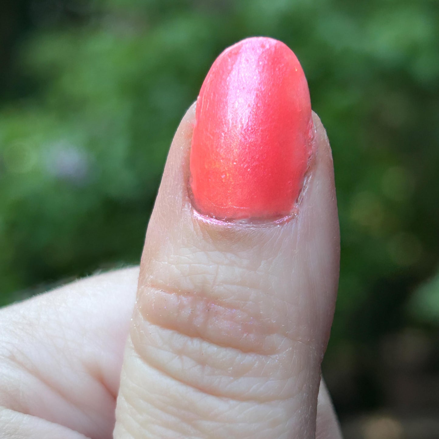 A thumb is shown outside painted in a bright neon pink-coral with shimmer.