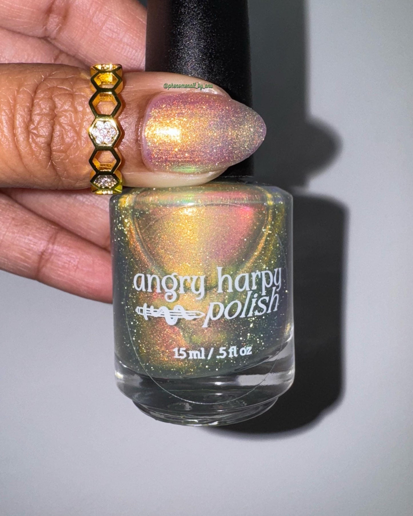 The scene is lit by flash. A ringed thumb is painted in a bright shifty polish that seems to glow gold-pink from within. An elegant bottle of the shifty polish with a white Angry Harpy Polish label is also shown.