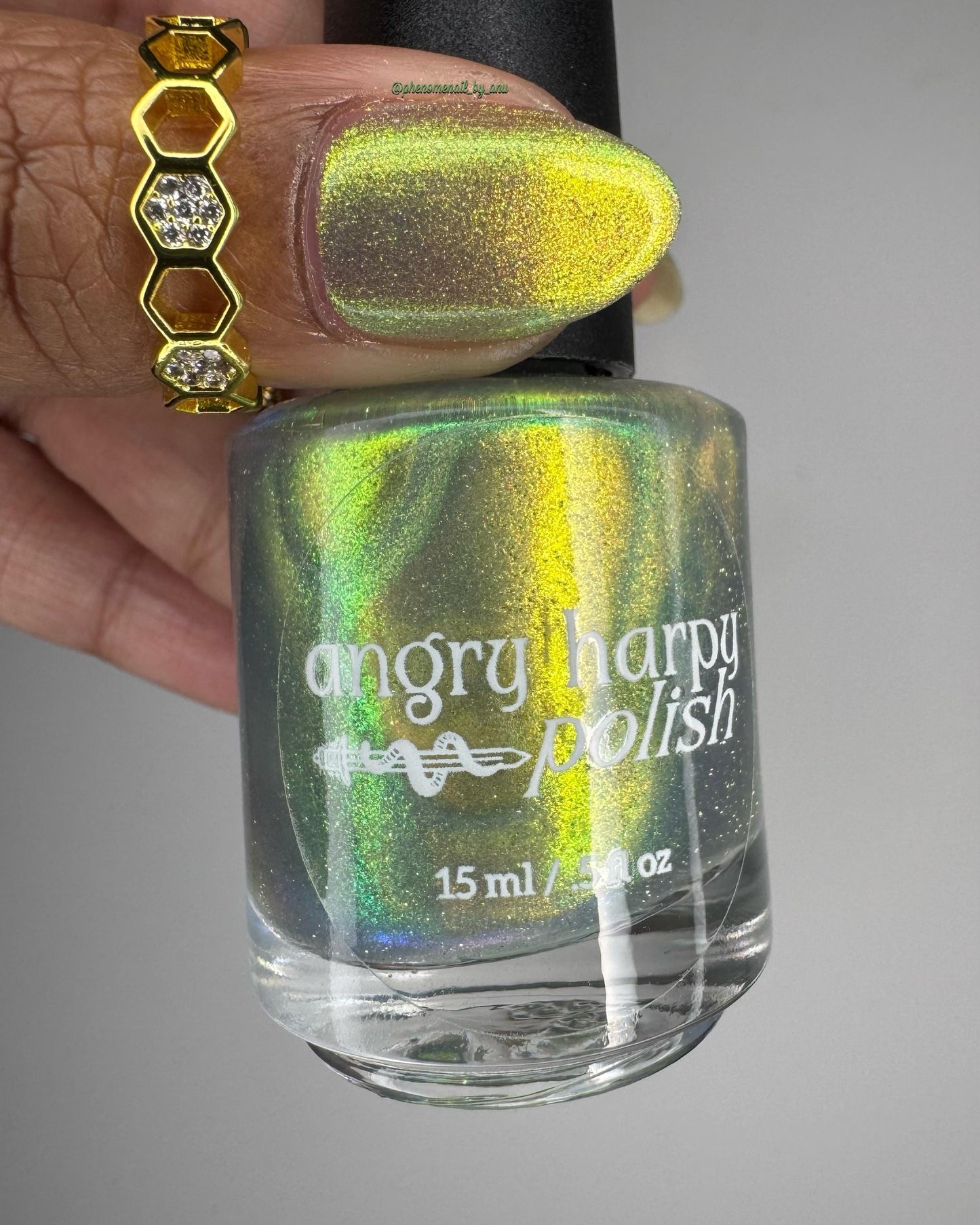 A ringed thumb is painted in a bright shifty polish that seems to glow yellow-green from within. An elegant bottle of the shifty polish with a white Angry Harpy Polish label is also shown.
