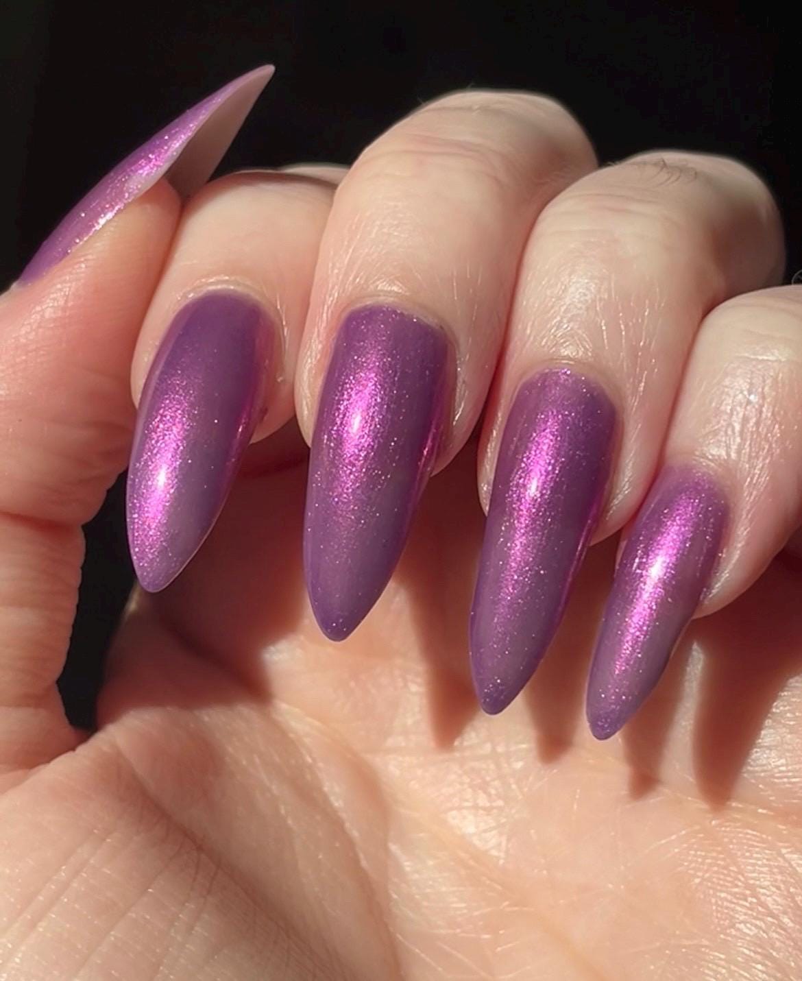 Long pale fingers end in long pointy nails painted with a medium purple shimmer nail polish. The polish has orange shimmer and scattered orange sparkle and glows in the light.