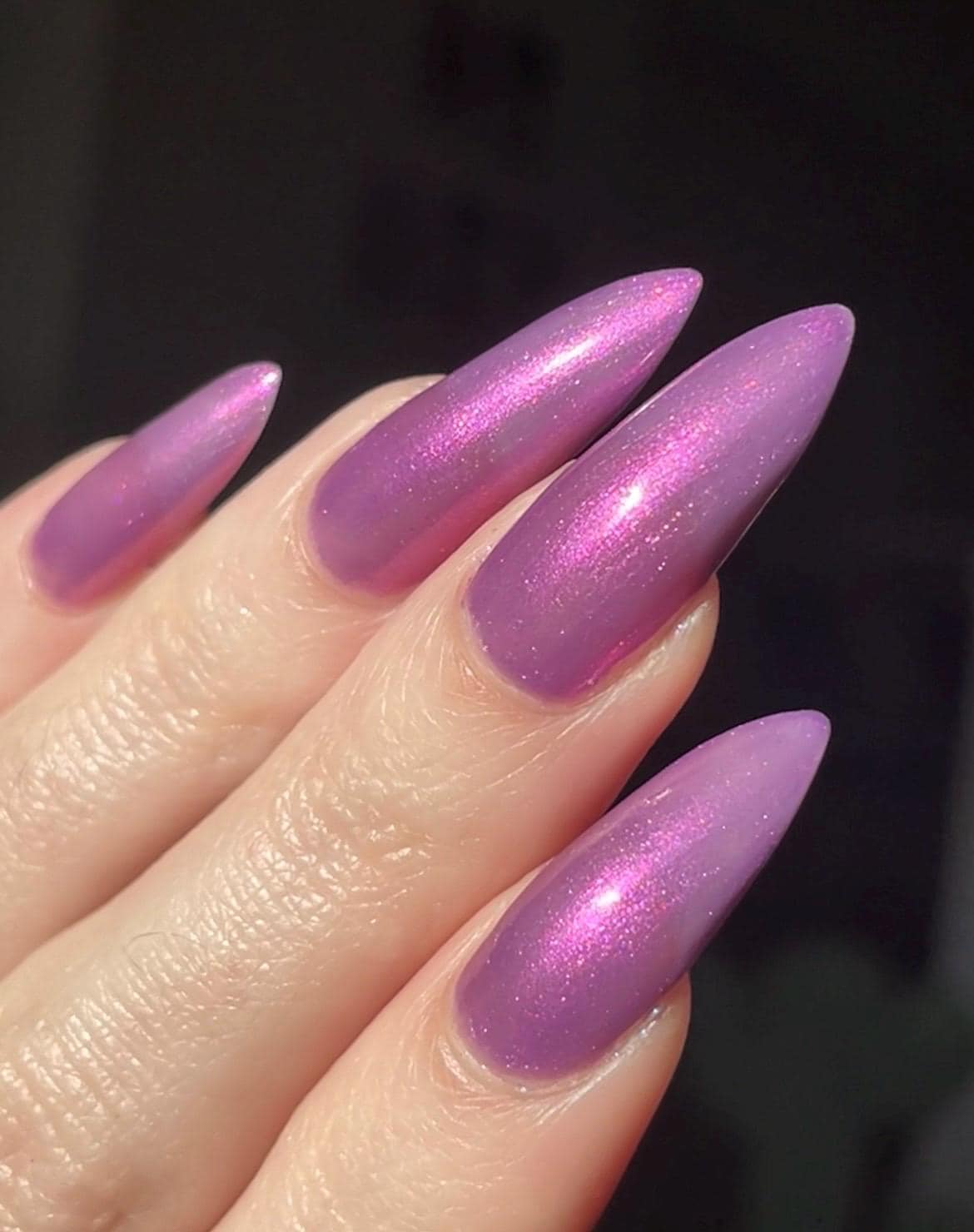 Long pale fingers end in long pointy nails painted with a medium purple shimmer nail polish. The polish has orange shimmer and scattered orange sparkle and glows in the light.