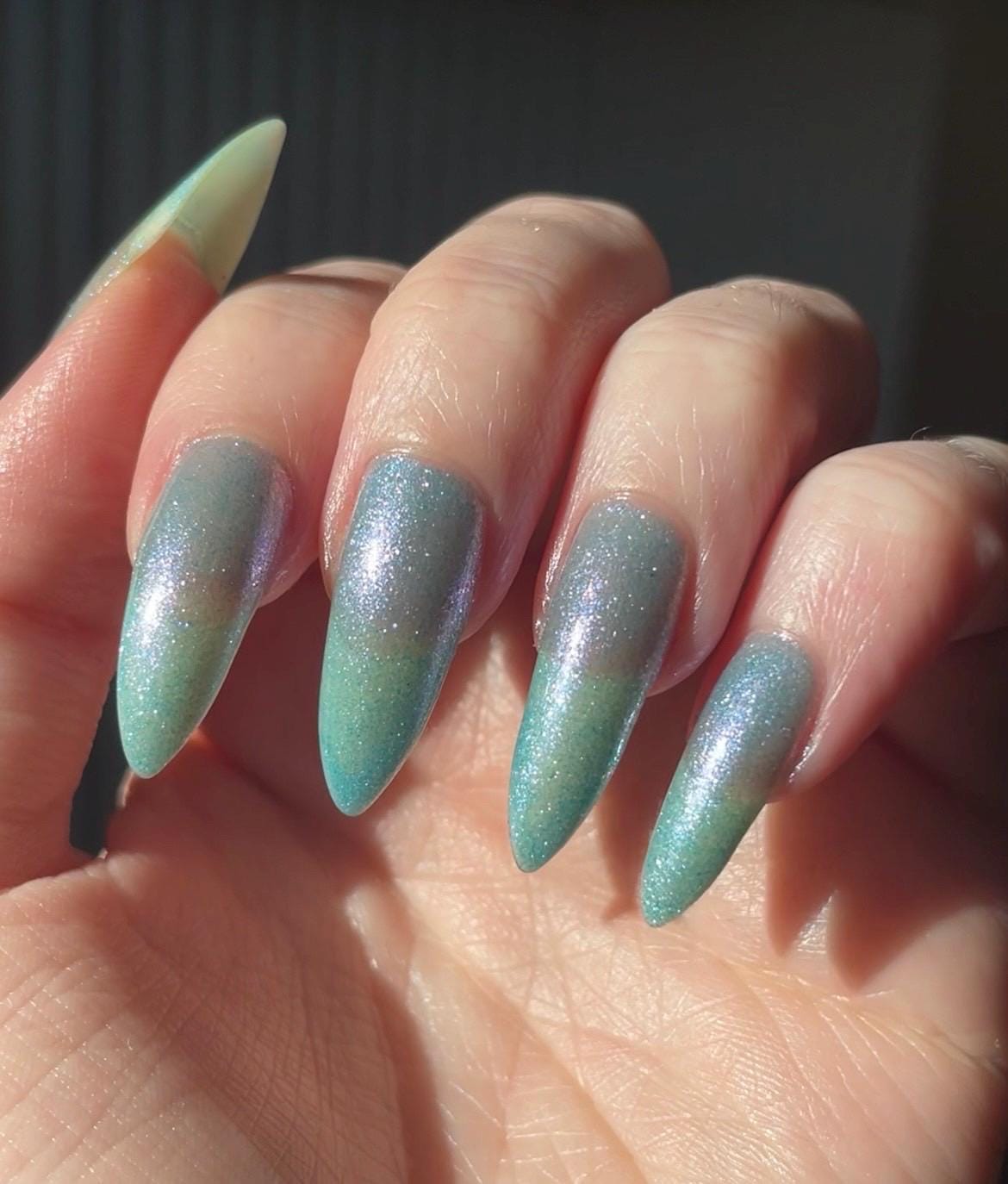 Long pale fingers end in long pointy nails painted with a pale blue shimmer nail polish. The polish has blue to pink shifting shimmer and glows in the light.