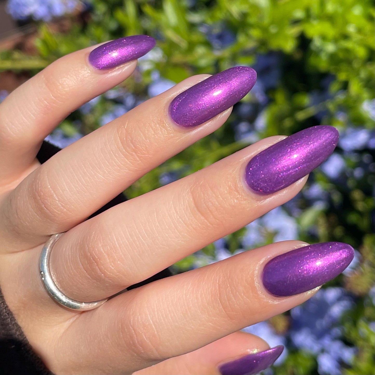 An elegant pale hand end in long oval nails painted with a medium purple shimmer nail polish. The polish has orange shimmer and scattered orange sparkle and glows in the outdoor light.