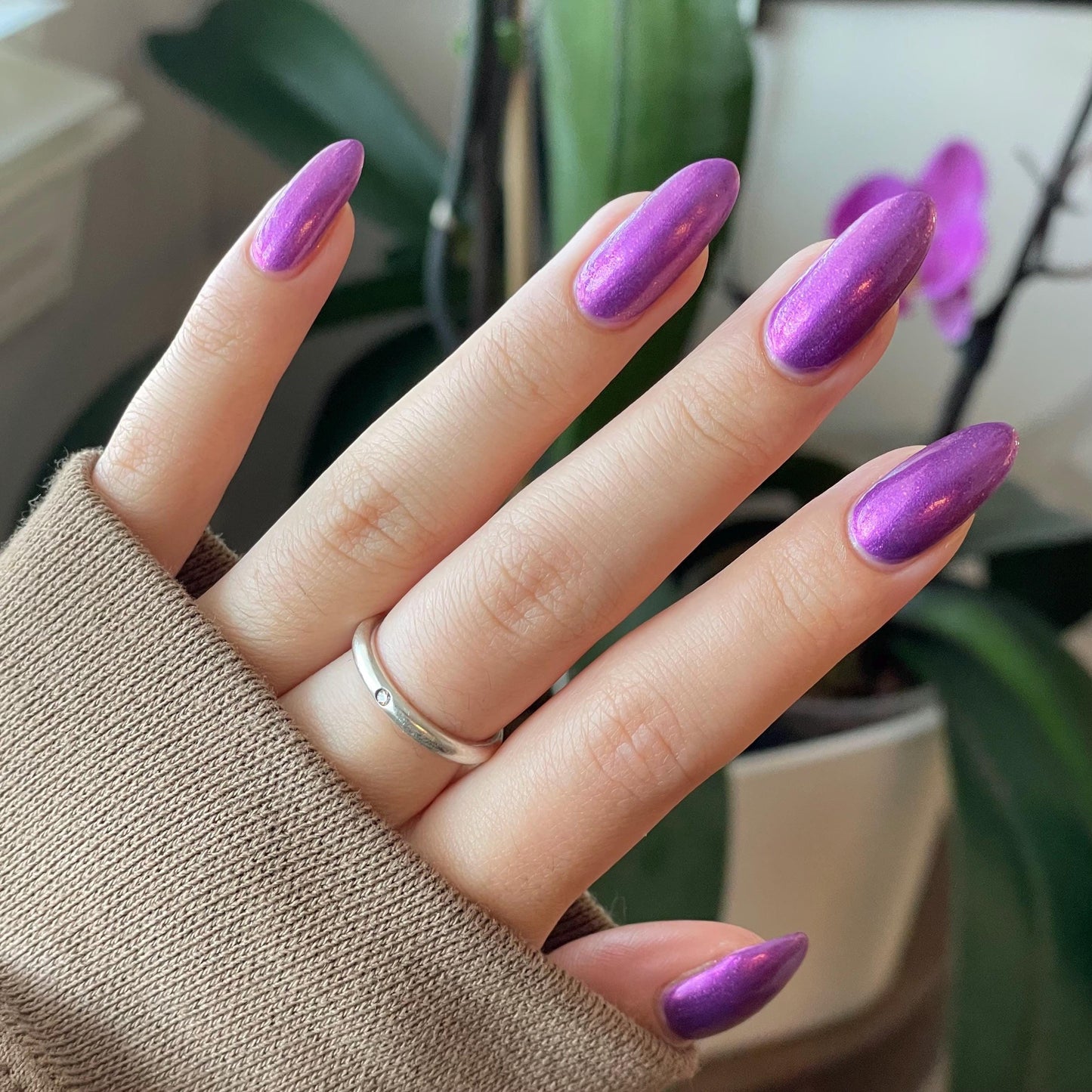 An elegant pale hand end in long oval nails painted with a medium purple shimmer nail polish. The polish has orange shimmer and scattered orange sparkle and glows in the indoor light.