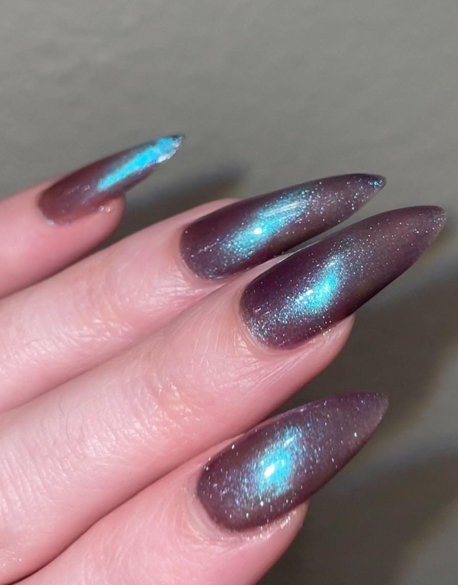 Long pointed nails are painted in a deep purple color that shimmers and sparkles with blue and purple magnetic particles. Magnetized in a stripe diagonally across the nails.