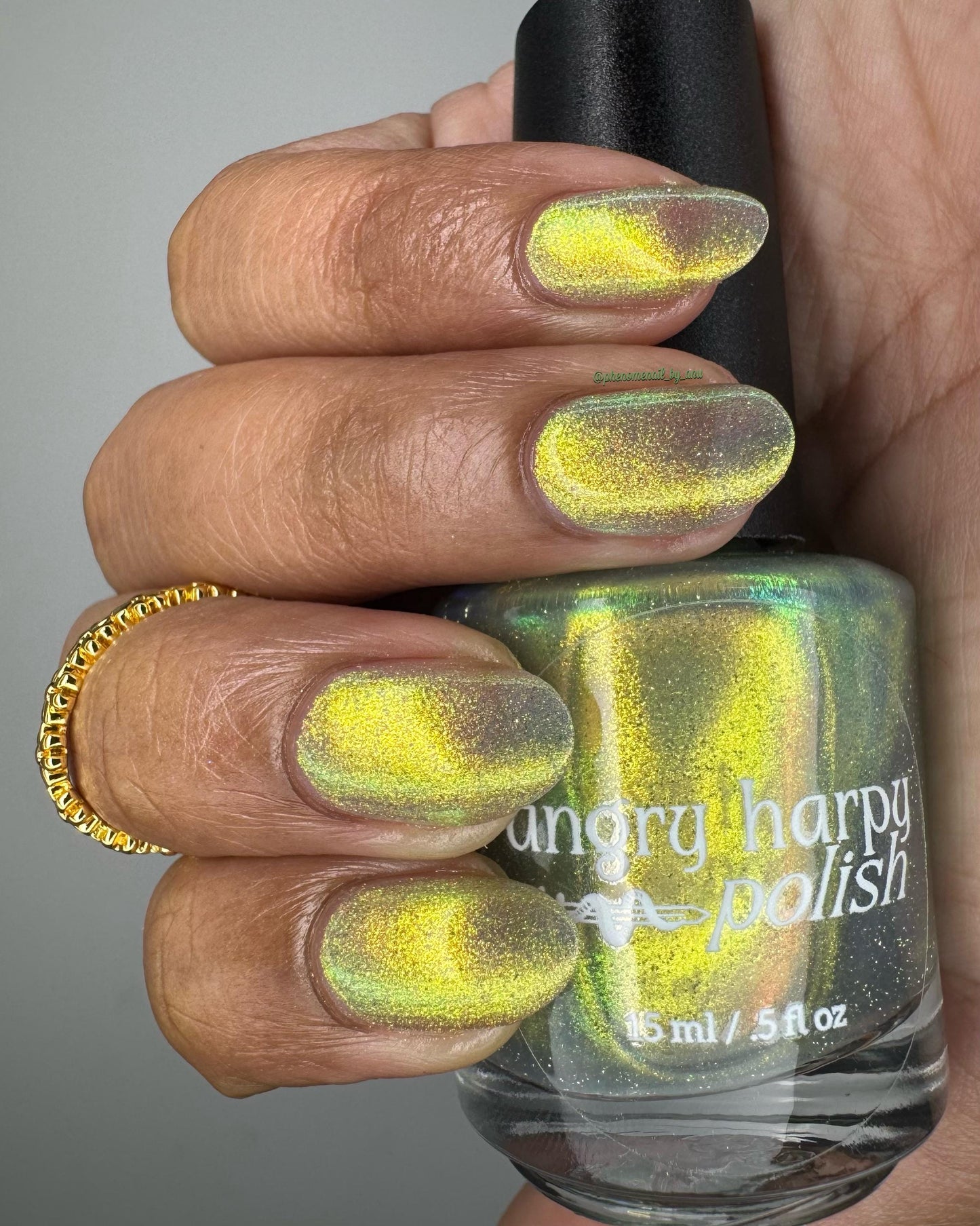 A hand with medium-dark skin holds a bottle of nail polish, showing off nails painted in a bright shifty polish that seems to glow yellow-green from within.