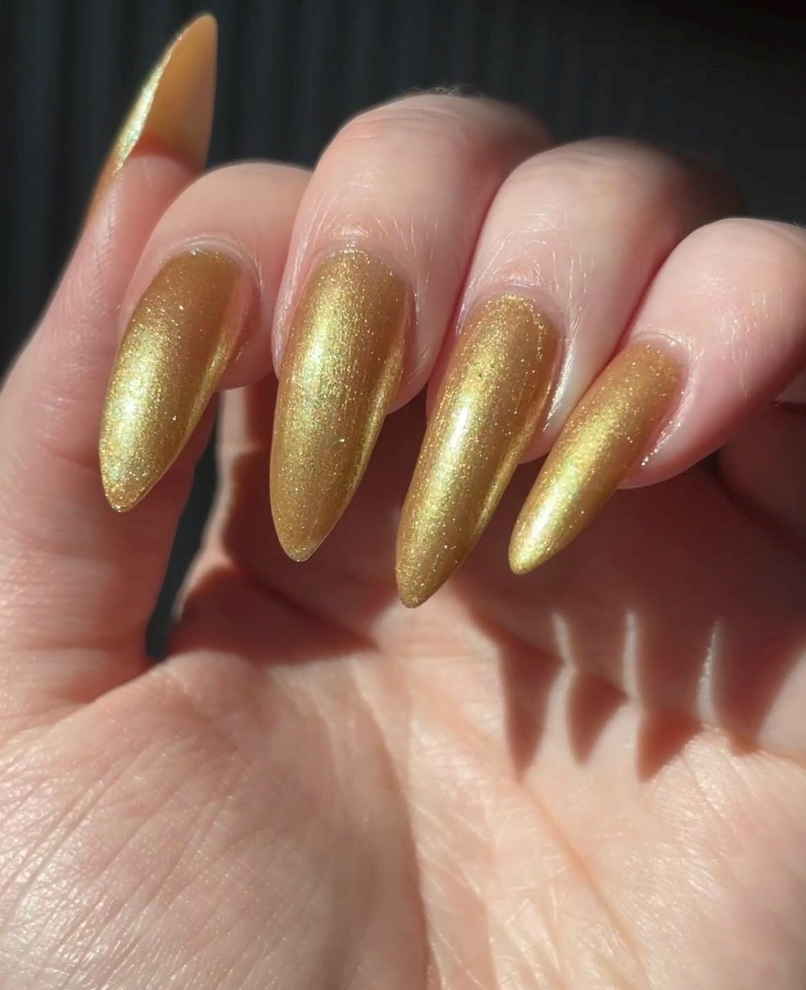 Long pale fingers end in long pointy nails painted with a golden shimmer nail polish. the polish has hints of green sparkle and glows in the light.