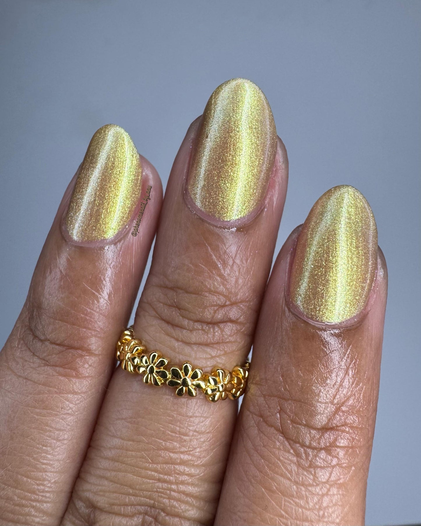 Three medium-brown fingers end in elegant oval nails painted with a golden shimmer nail polish. The polish has hints of green sparkle and glows in the light.