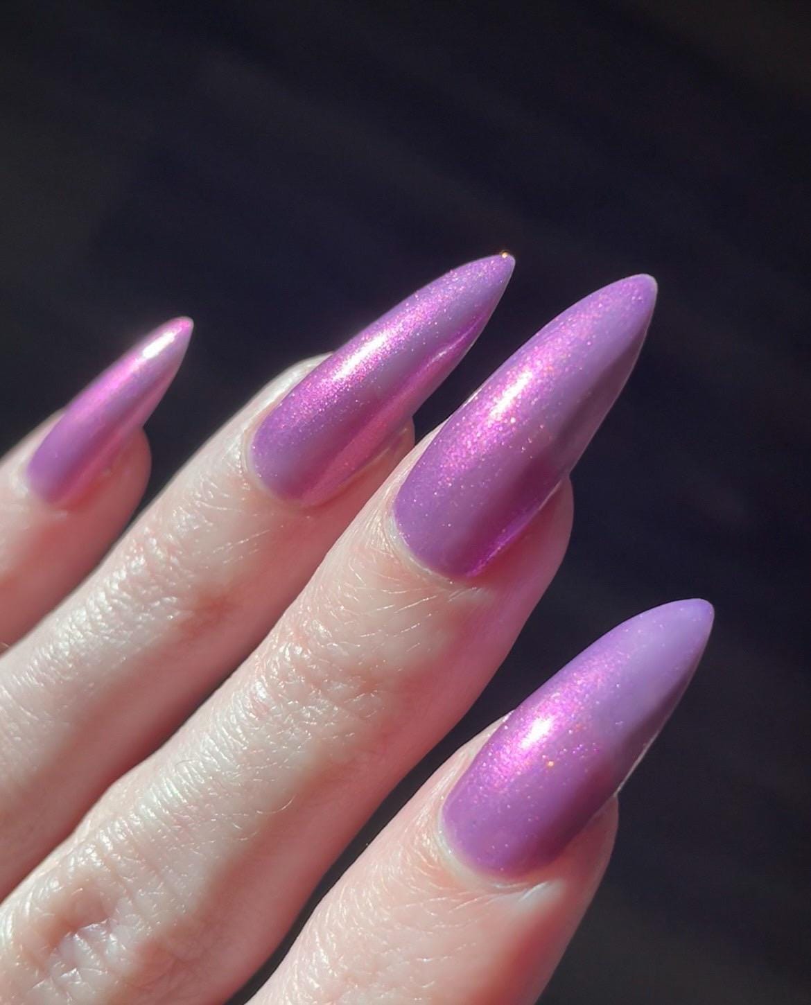 Long pale fingers end in long pointy nails painted with a medium purple shimmer nail polish. The polish has orange shimmer and scattered orange sparkle and glows in the light.