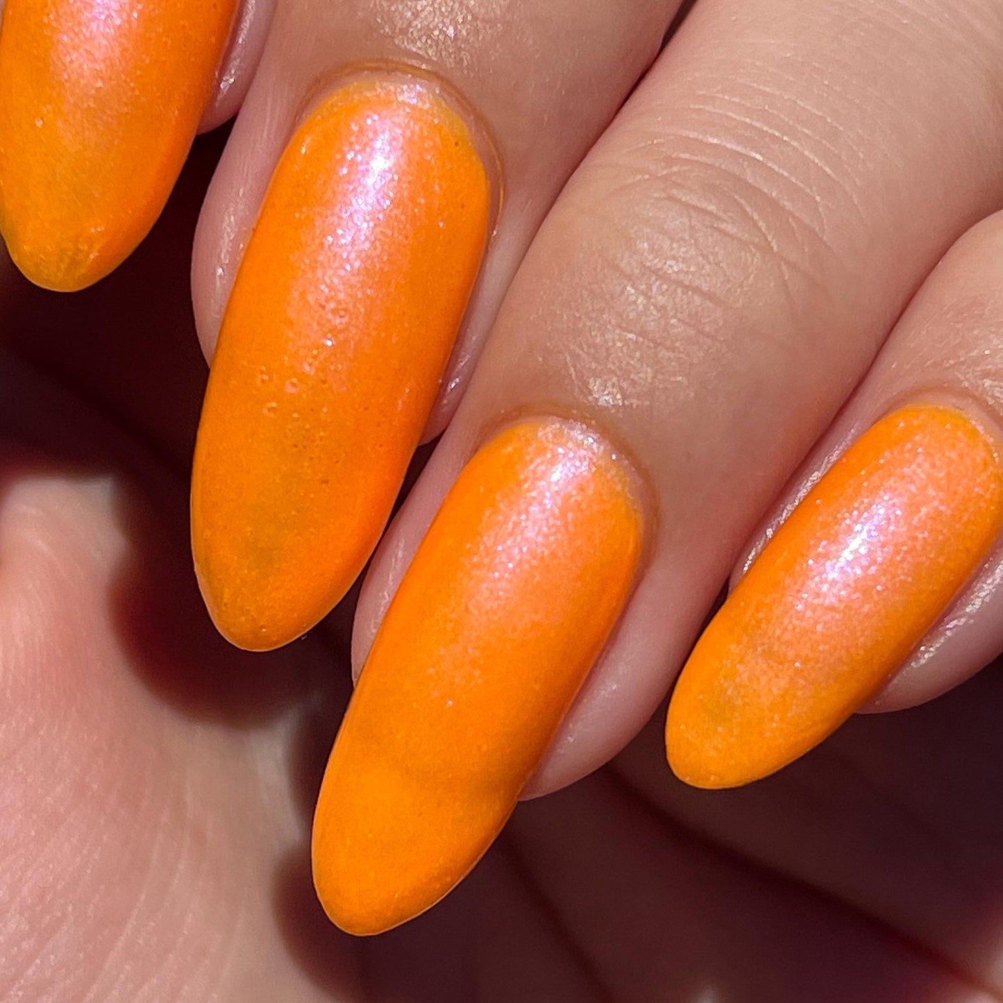 A closeup of a hand with nails painted neon orange nail lacquer with pink-purple shimmer.