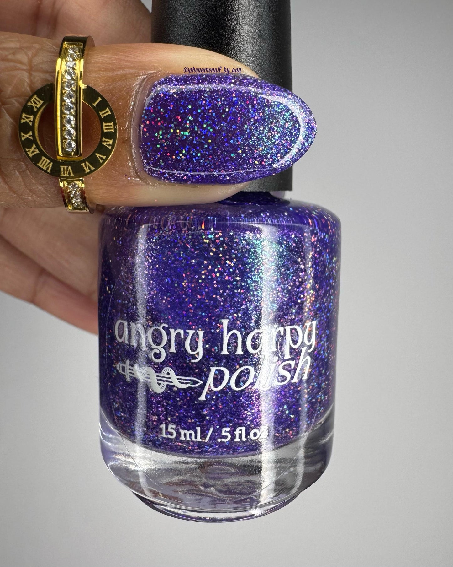 A closeup of a thumb and nail polish bottle with a vibrant violet lacquer sparkling with purple holographic glitter and green shimmer.