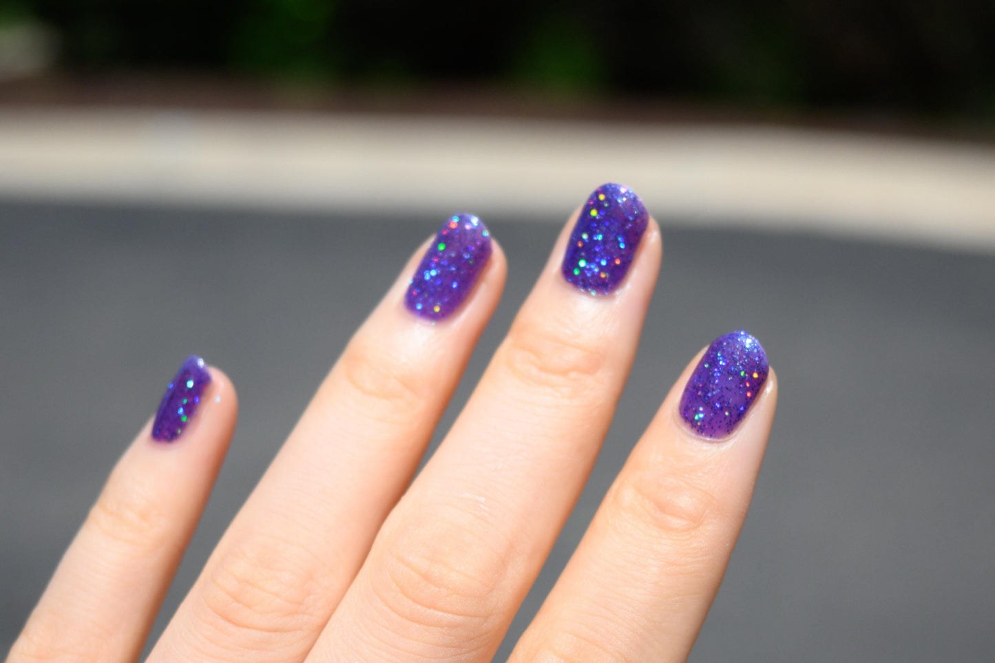 A closeup of a hand with nails painted a vibrant violet lacquer sparkling with purple holographic glitter and green shimmer.