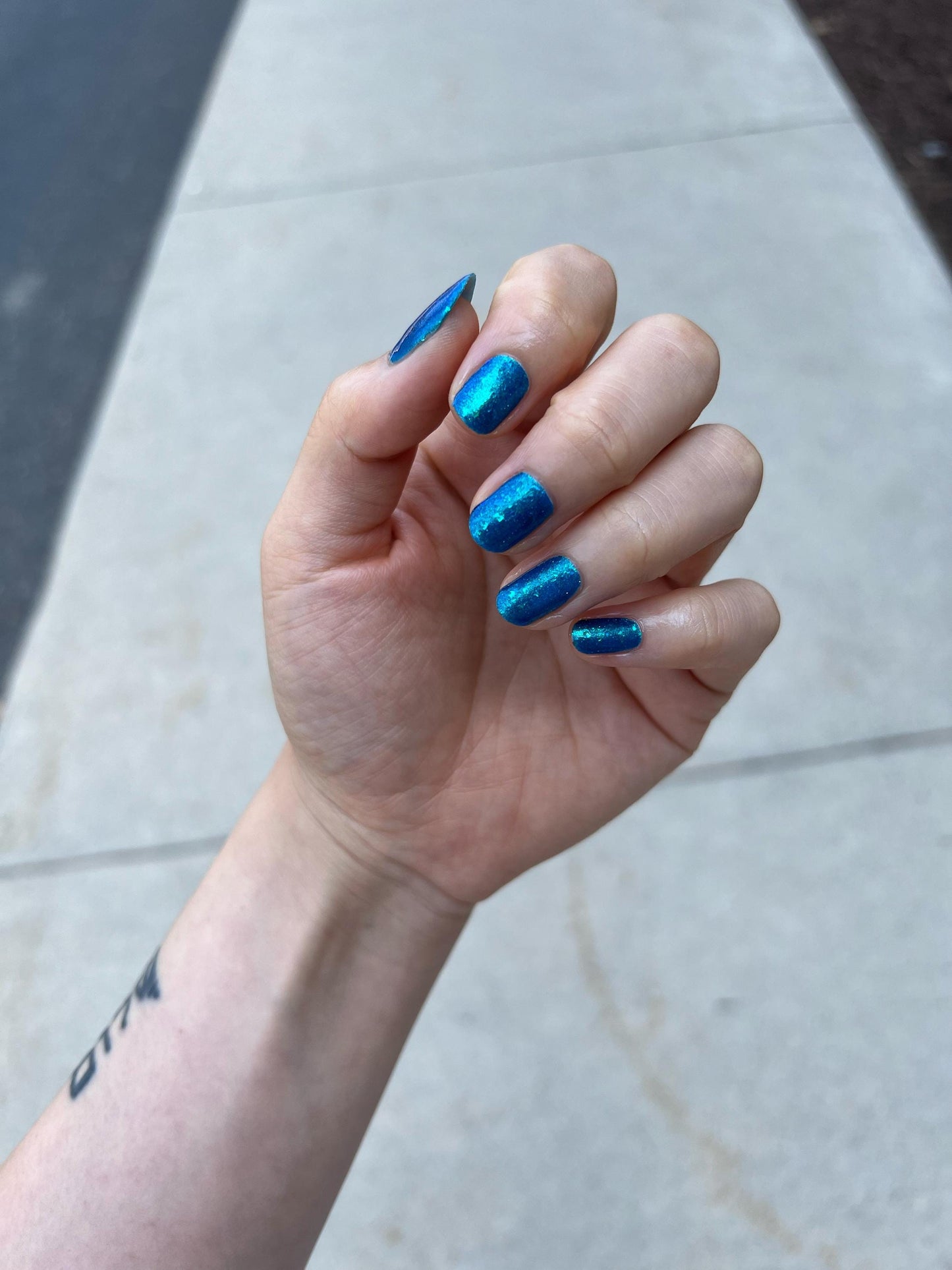 A closeup of a hand with nails painted a gorgeous, vibrant, shimmery deep teal color, packed with flakes and reflective blue glitter scattered throughout.