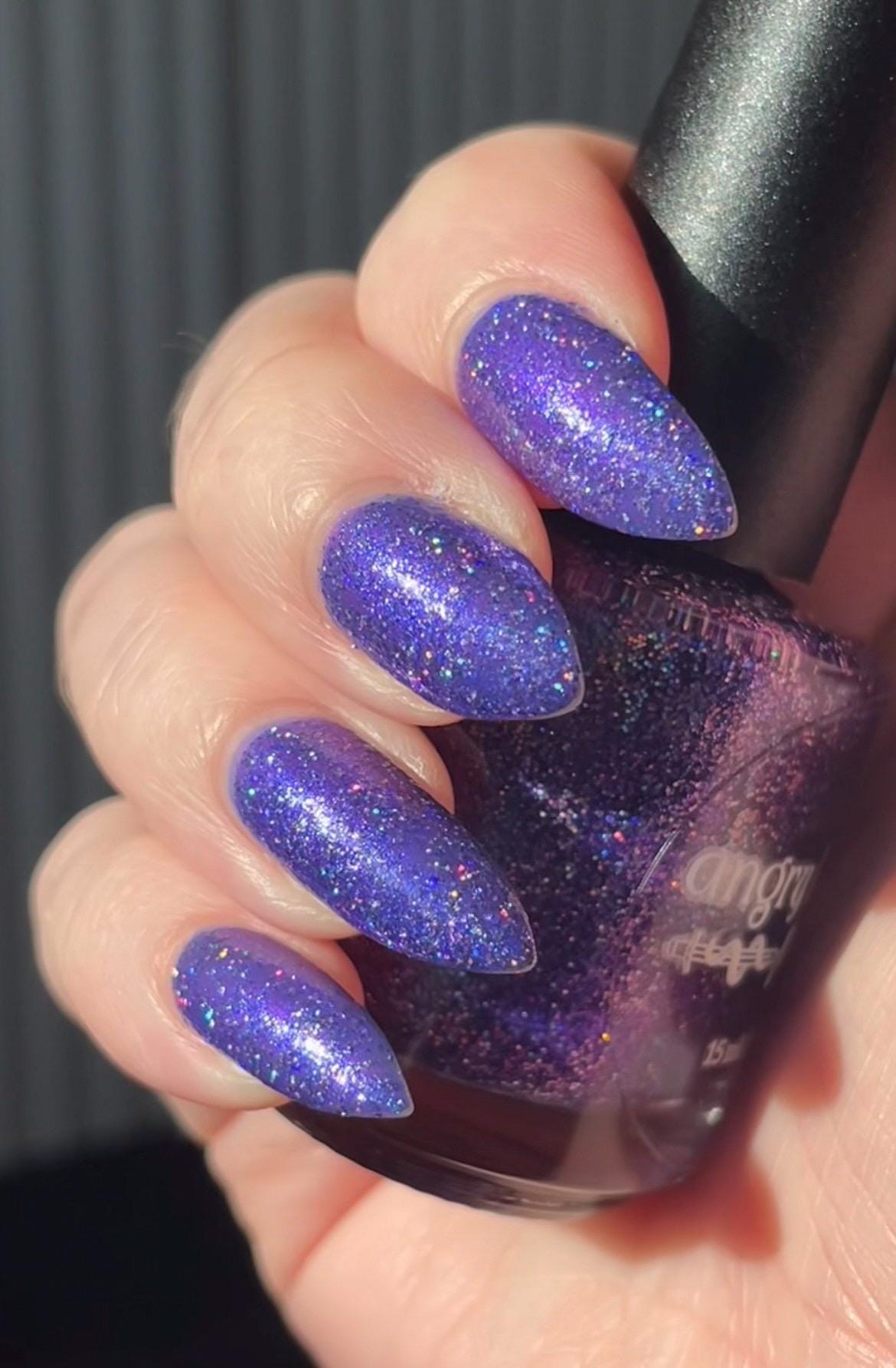 A closeup of a hand with nails painted a vibrant violet lacquer sparkling with purple holographic glitter and green shimmer.