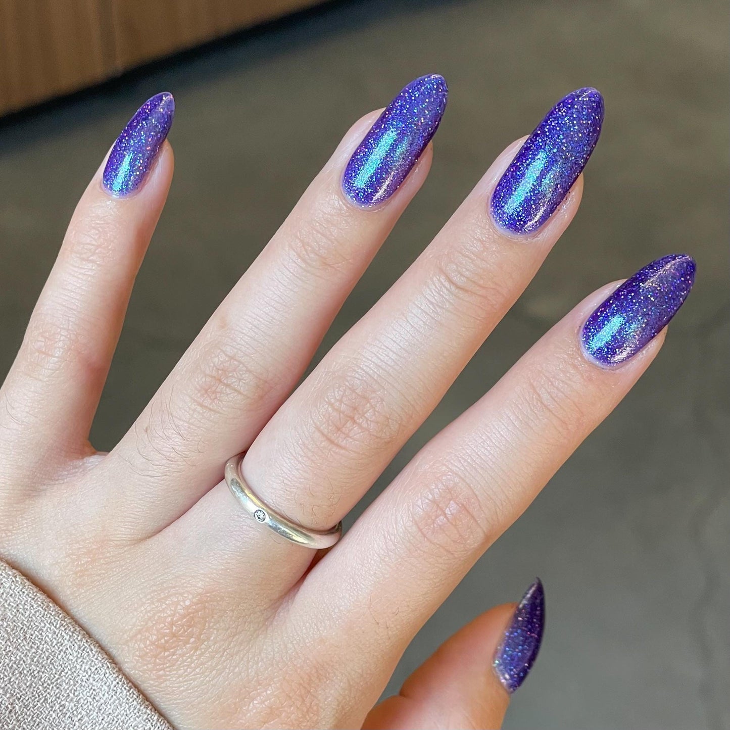 A closeup of a hand with nails painted a vibrant violet lacquer sparkling with purple holographic glitter and green shimmer.