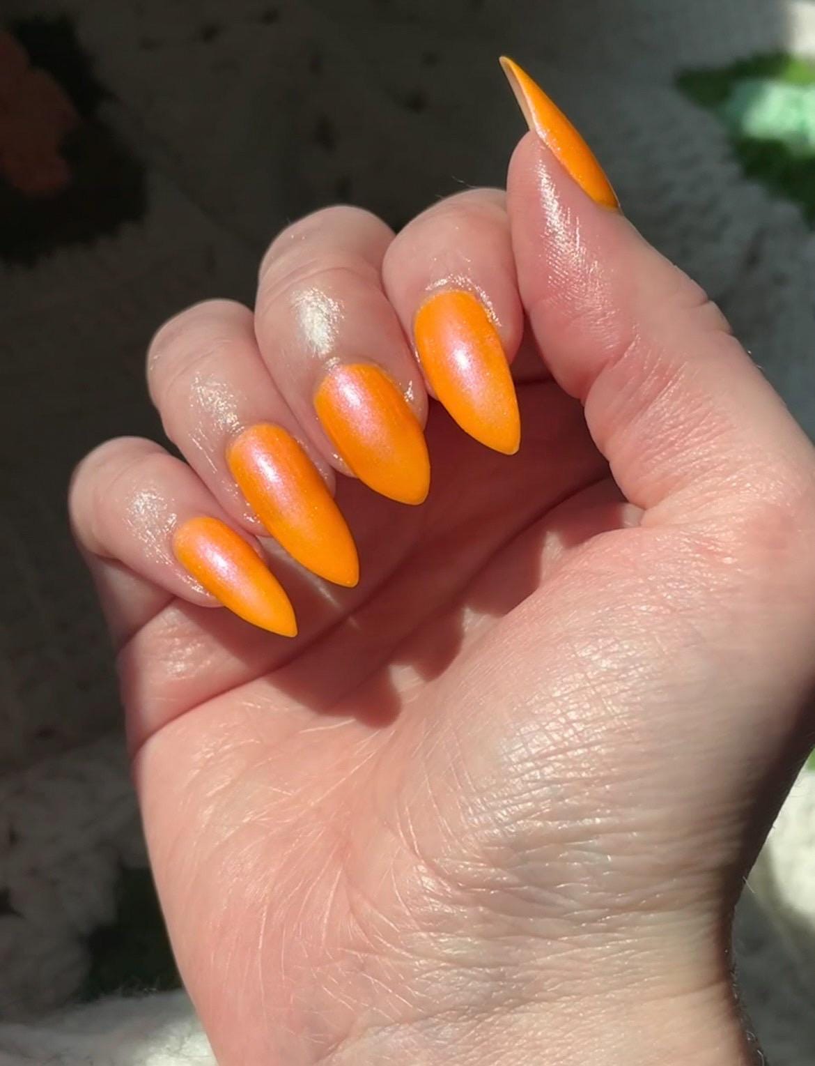 A closeup of a hand with nails painted neon orange nail lacquer with pink-purple shimmer.