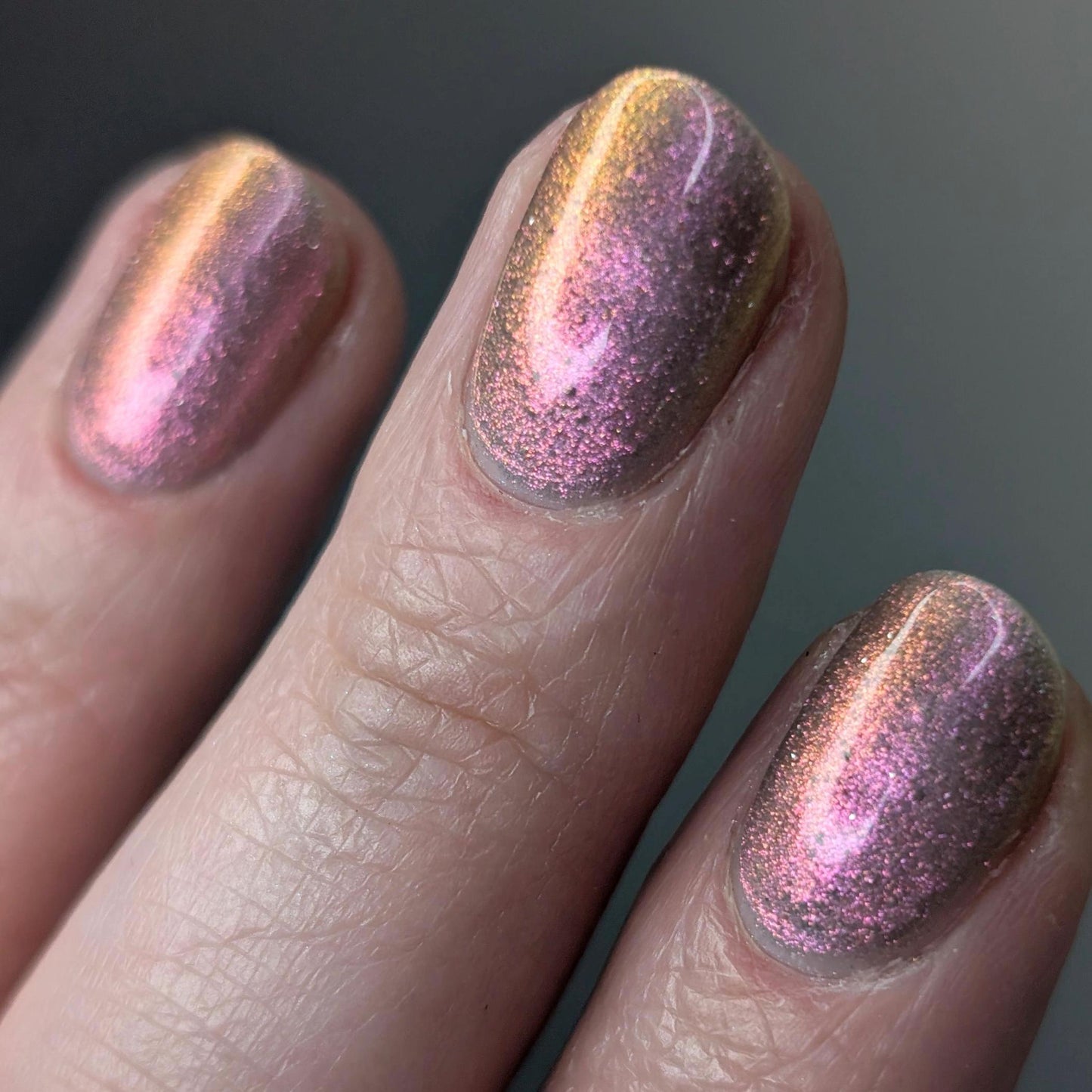 A closeup of a hand with nails painted a sheer multichrome nail polish that shifts gold-orange-pink-fuschia, with pale pink reflective glitter scattered throughout.