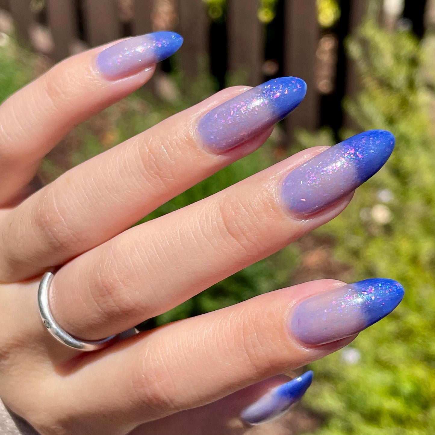 A closeup of a hand with nails painted a thermal polish that goes from dark blue at the tips (cold) to a blue-tinted clear (warm). Polish is filled with iridescent green-pink flakes and blue shimmer.