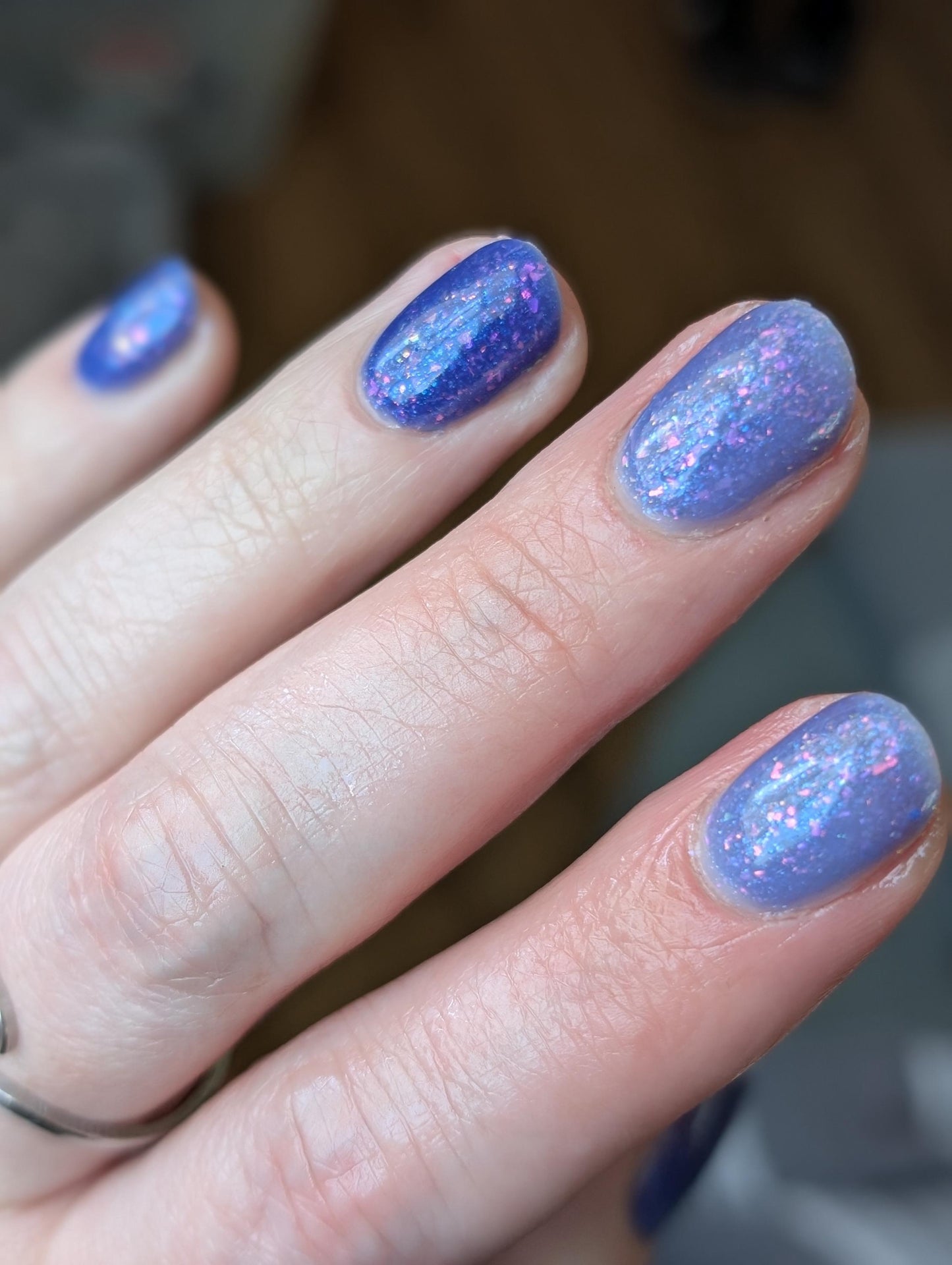 A closeup of a hand with nails painted a thermal polish that goes from dark blue at the tips (cold) to a blue-tinted clear (warm). Polish is filled with iridescent green-pink flakes and blue shimmer.