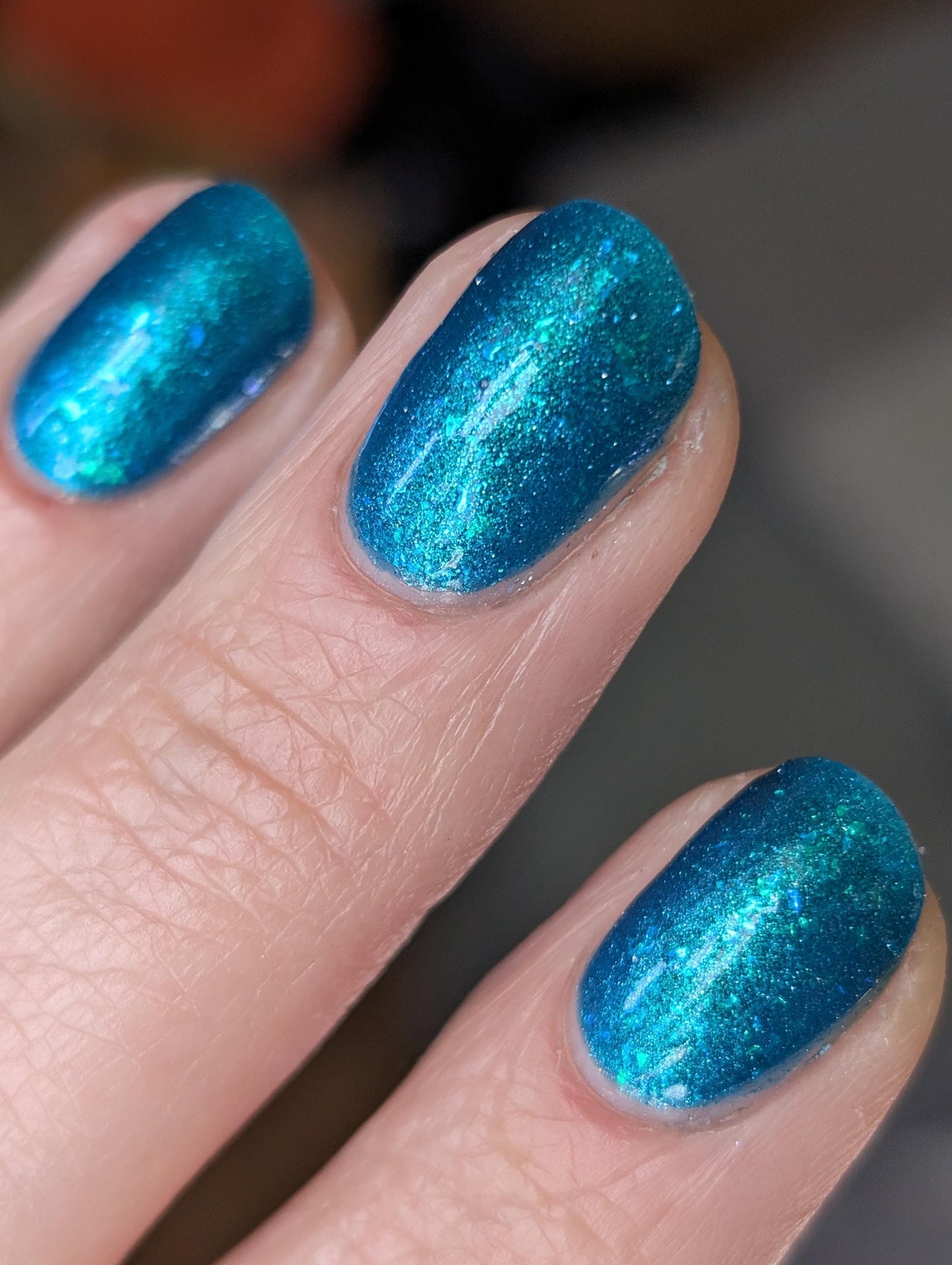 A closeup of a hand with nails painted a gorgeous, vibrant, shimmery deep teal color, packed with flakes and reflective blue glitter scattered throughout.
