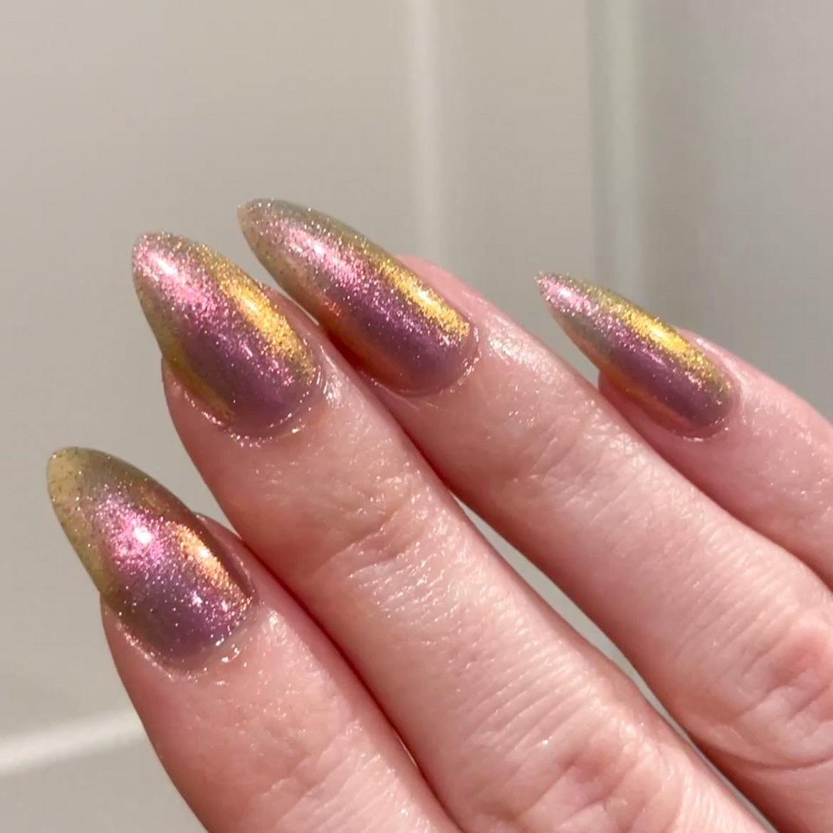 A closeup of a hand with nails painted a sheer multichrome nail polish that shifts gold-orange-pink-fuschia, with pale pink reflective glitter scattered throughout.