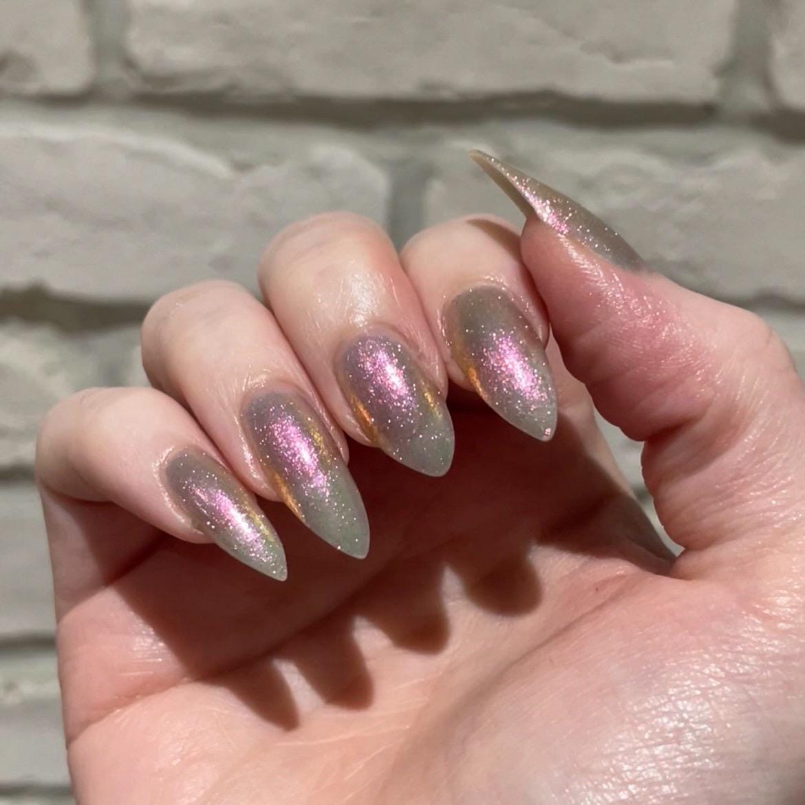 A closeup of a hand with nails painted a sheer multichrome nail polish that shifts gold-orange-pink-fuschia, with pale pink reflective glitter scattered throughout.