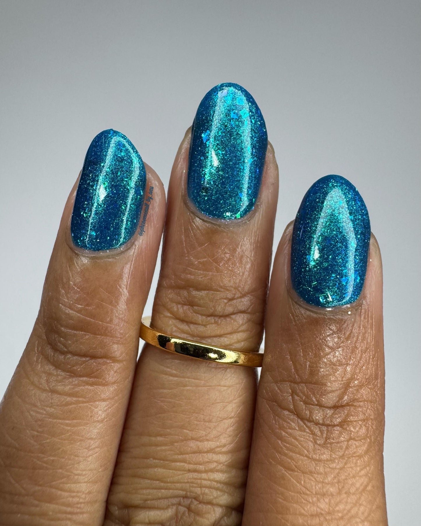 A closeup of a hand with nails painted a gorgeous, vibrant, shimmery deep teal color, packed with flakes and reflective blue glitter scattered throughout.