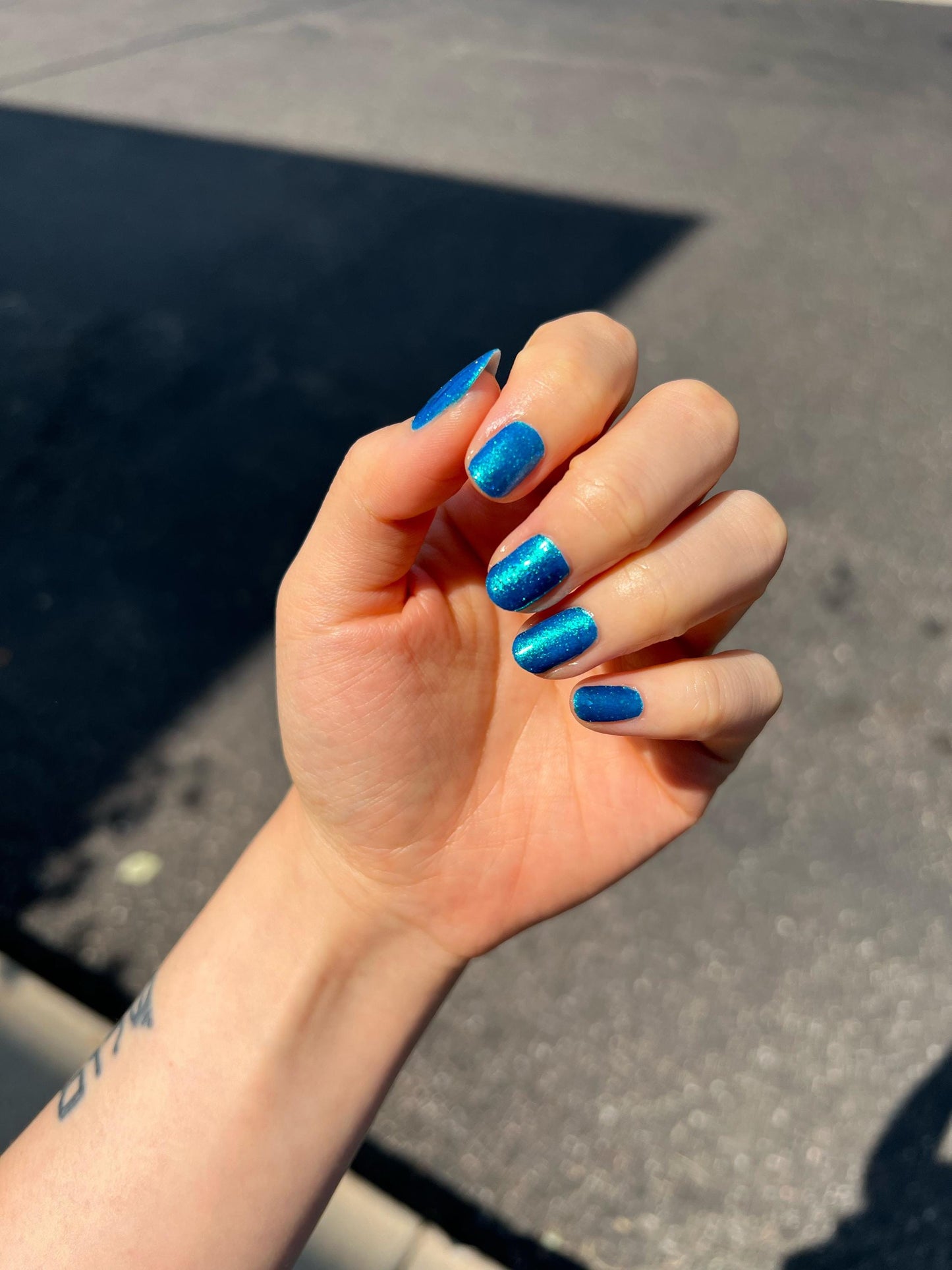 A closeup of a hand with nails painted a gorgeous, vibrant, shimmery deep teal color, packed with flakes and reflective blue glitter scattered throughout.