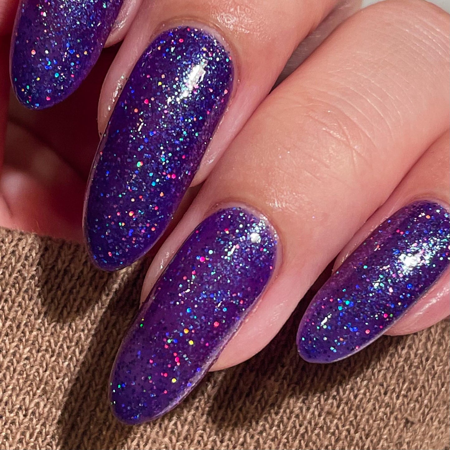 A closeup of a hand with nails painted a vibrant violet lacquer sparkling with purple holographic glitter and green shimmer.