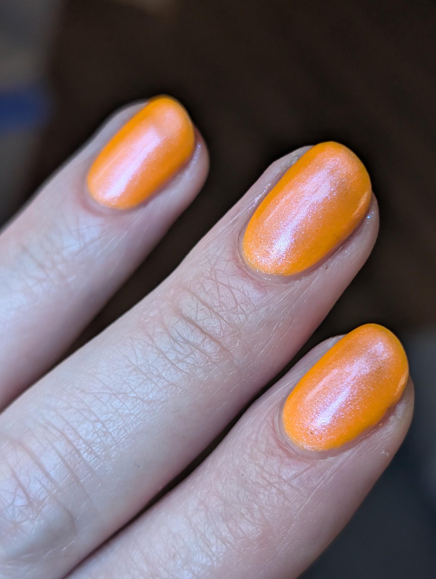 A closeup of a hand with nails painted neon orange nail lacquer with pink-purple shimmer.