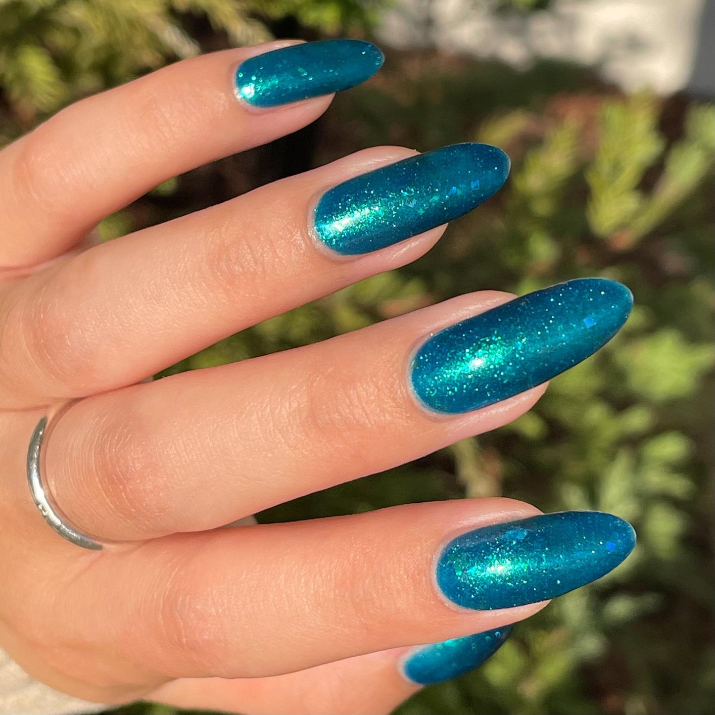 A closeup of a pale hand with nails painted a gorgeous, vibrant, shimmery deep teal color, packed with flakes and reflective blue glitter scattered throughout.
