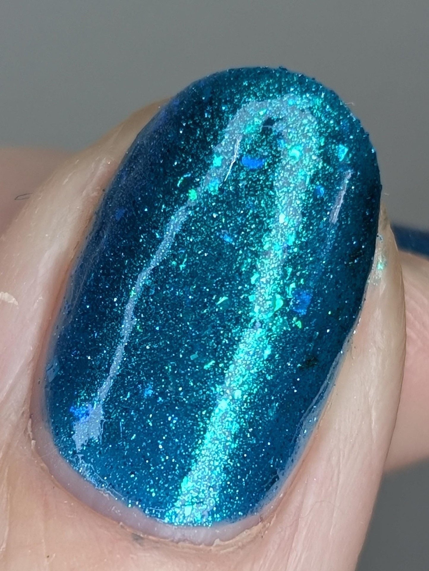 A closeup of a hand with nails painted a gorgeous, vibrant, shimmery deep teal color, packed with flakes and reflective blue glitter scattered throughout.