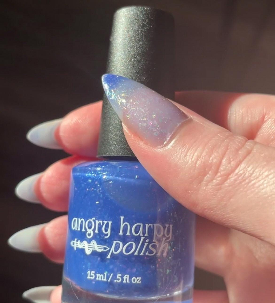 A closeup of a hand with nails painted a thermal polish that goes from dark blue at the tips (cold) to a blue-tinted clear (warm). Polish is filled with iridescent green-pink flakes and blue shimmer.