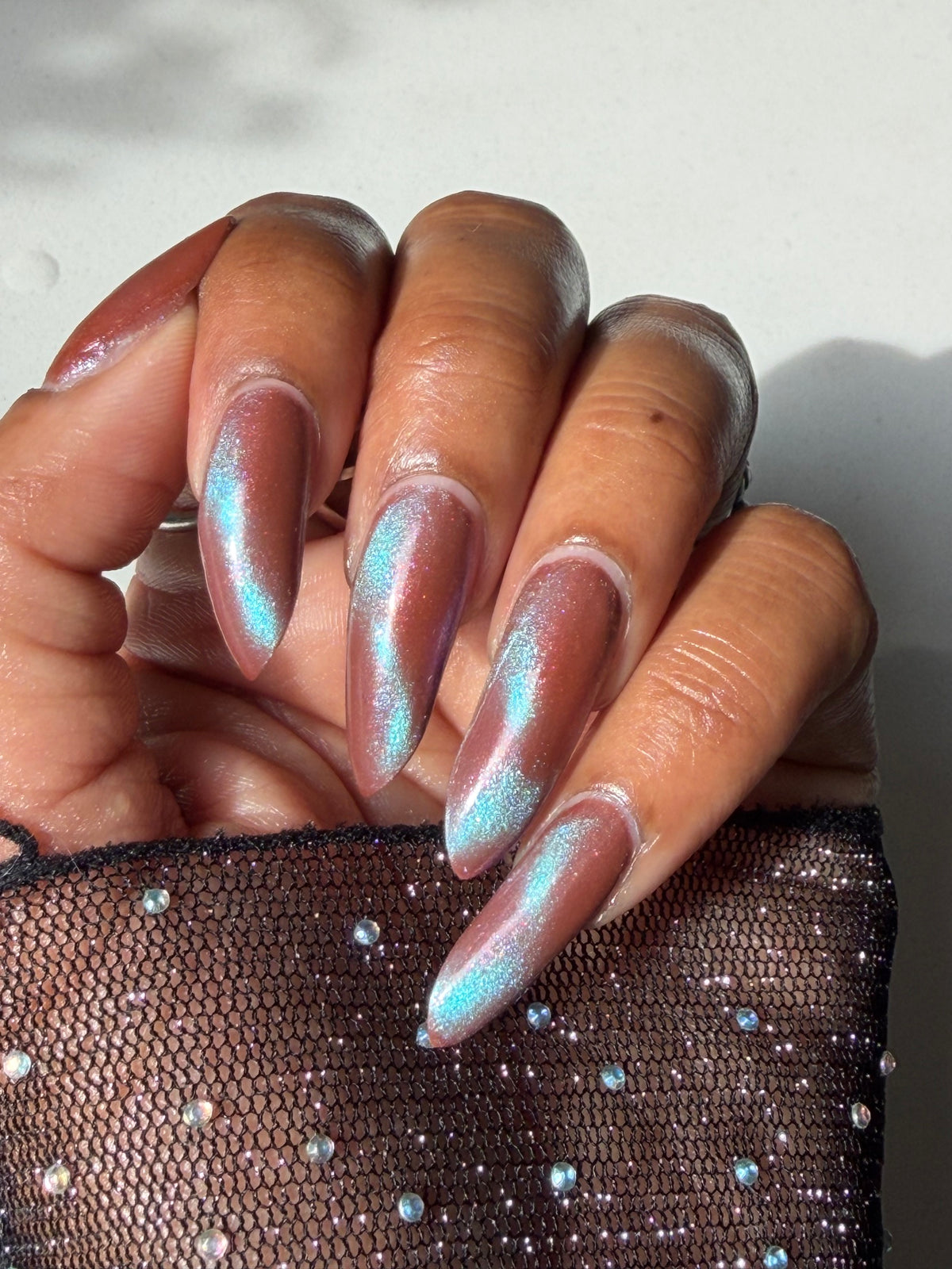 A close-up of a hand with long stiletto-shaped nails painted in a shimmery iridescent pink and light blue holographic finish, next to a sheer black mesh sleeve with rhinestones.