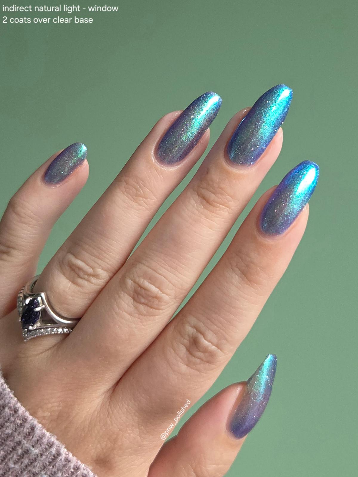 A close-up of a hand with long, almond-shaped nails painted in a shimmering iridescent nail polish that shifts between blue, teal, and purple. The hand is shown against a solid sage green background, wearing a silver ring on the left hand.