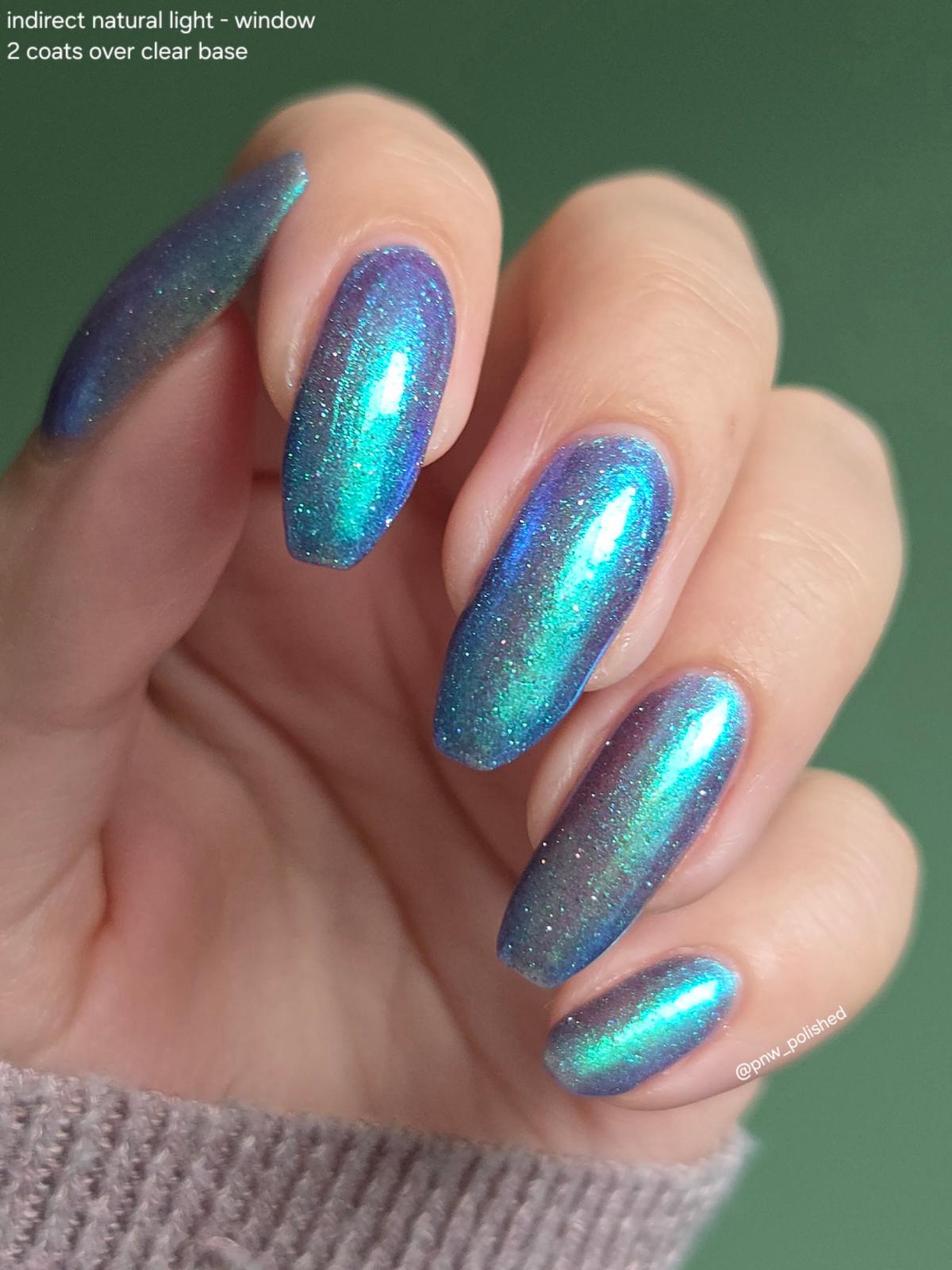 A close-up of a hand with long, almond-shaped nails painted in a shimmery iridescent blue-to-green holographic nail polish. Text in the top-left corner reads 'indirect natural light - window 2 coats over clear base', and a small watermark is visible on the bottom-right.