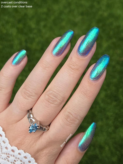 A close-up of a hand with long almond-shaped nails painted in an iridescent holographic nail polish that shifts between teal, blue, and purple under overcast light. The hand is wearing a silver ring with a blue stone, set against a blurred green grass background.