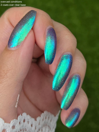 A close-up of a hand with long almond-shaped nails painted in a shimmering holographic nail polish that shifts between teal, green, blue, and purple, set against a blurred green background. Text in the top left reads 'overcast conditions 2 coats over clear base'.