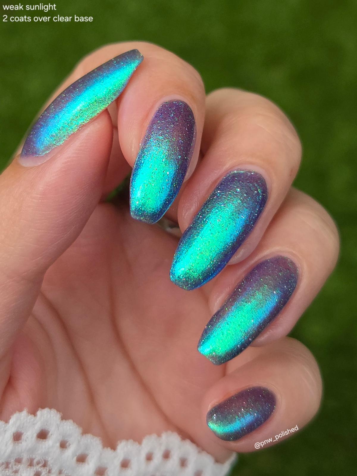 A close-up of a hand with long almond-shaped nails painted in an iridescent holographic nail polish that shifts between teal, blue, and purple, with glitter. The background is a soft-focus green outdoor setting.