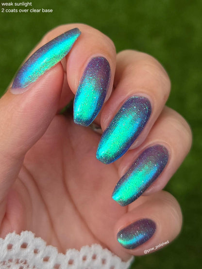 A close-up of a hand with long almond-shaped nails painted in an iridescent holographic nail polish that shifts between teal, blue, and purple, with glitter. The background is a soft-focus green outdoor setting.