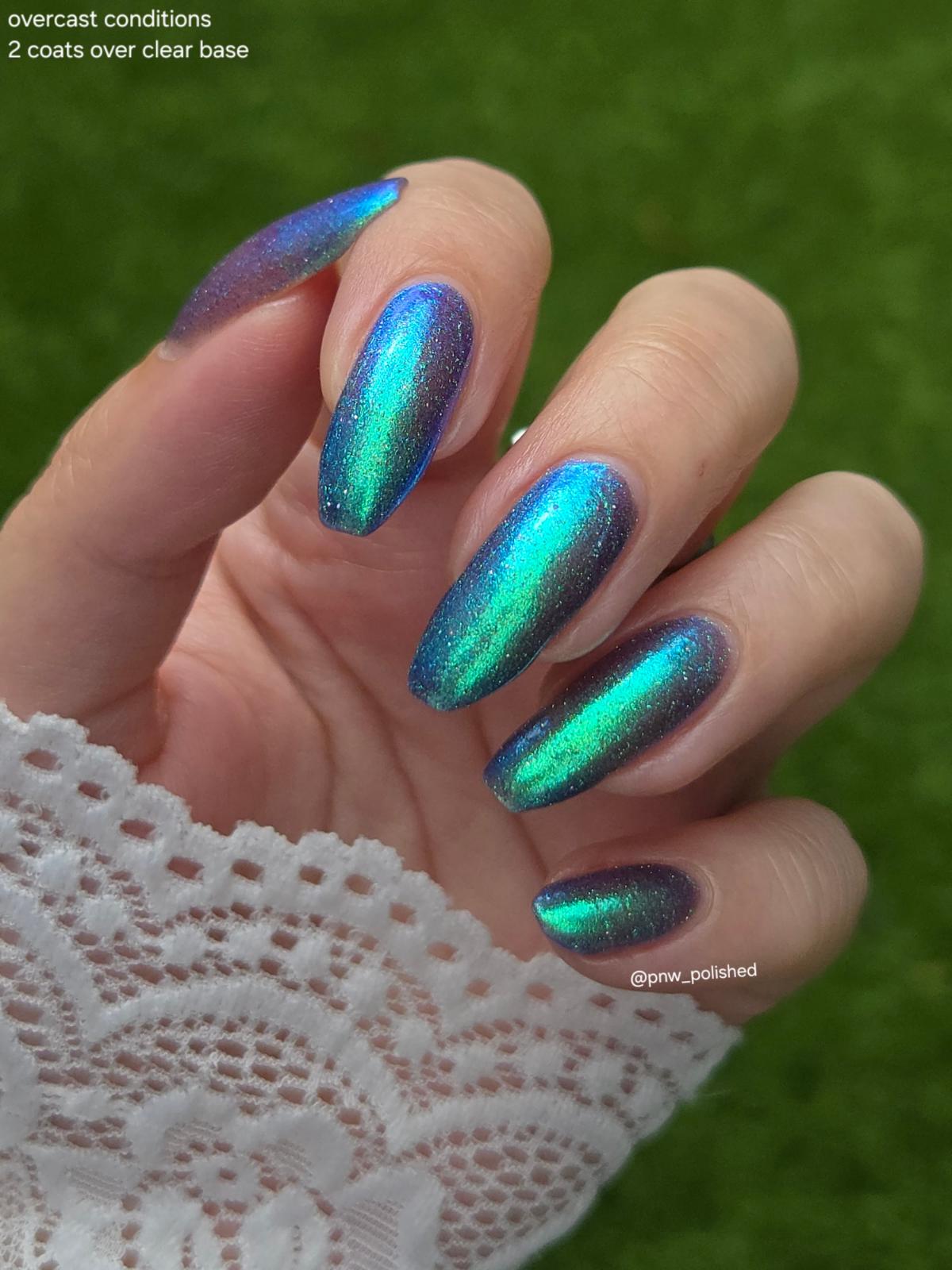 A close-up of a hand with long almond-shaped nails painted in a holographic iridescent nail polish that shifts between teal, blue, and purple, set against a blurred green background. White lace is visible at the wrist, and text in the top-left corner reads 'overcast conditions 2 coats over clear base'.