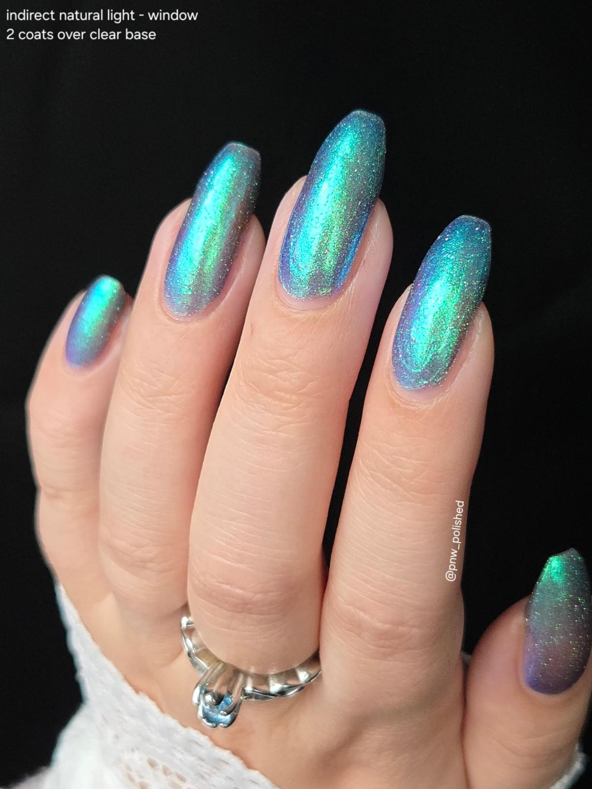 A close-up of a hand with long almond-shaped nails painted in a holographic teal and blue shimmer nail polish, shown against a black background with text noting indirect natural light and 2 coats over clear base.