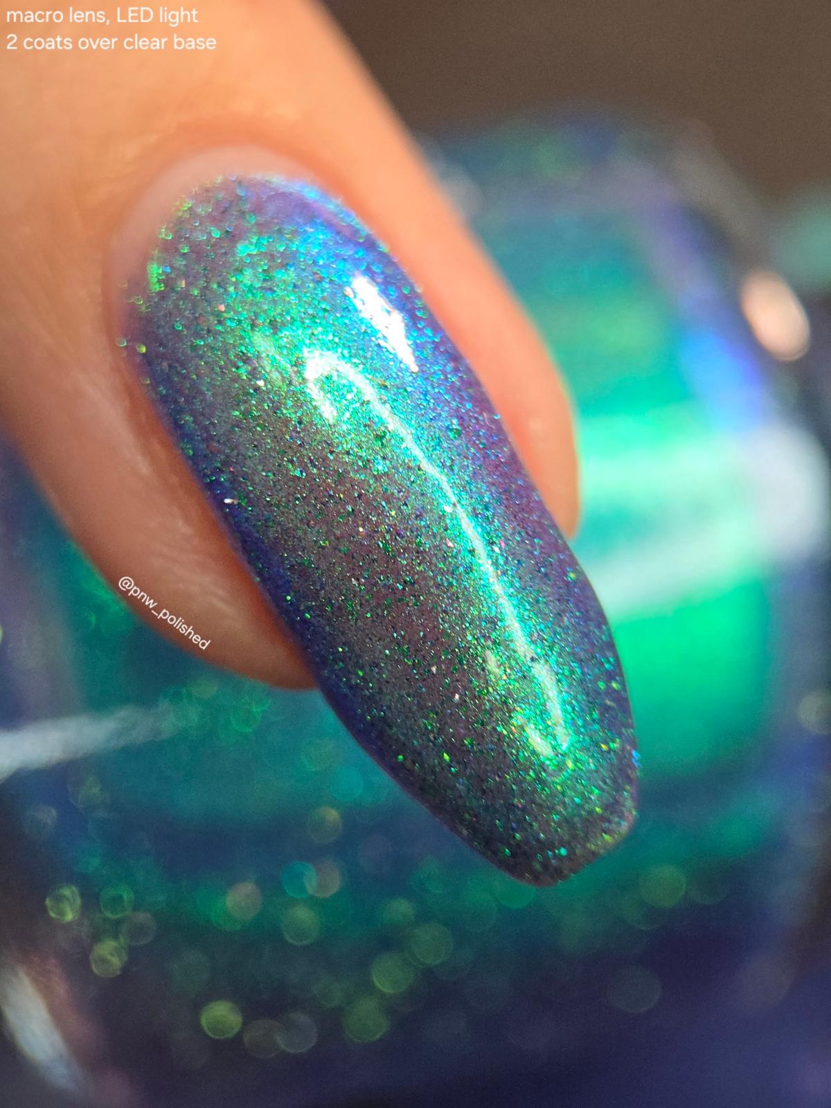 A close-up of a fingernail painted with a holographic glitter nail polish that shifts between teal, green, blue, and purple depending on the light. The text in the top-left corner reads 'macro lens, LED light 2 coats over clear base'.