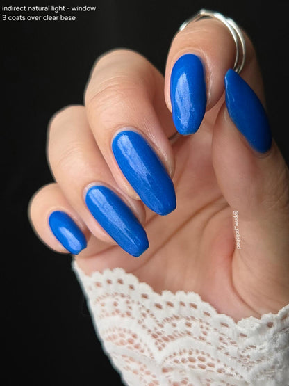 A close-up of a hand with long, almond-shaped nails painted in a glossy, vibrant medium blue. The nails are shown against a solid black background, with a white lace sleeve visible at the wrist.