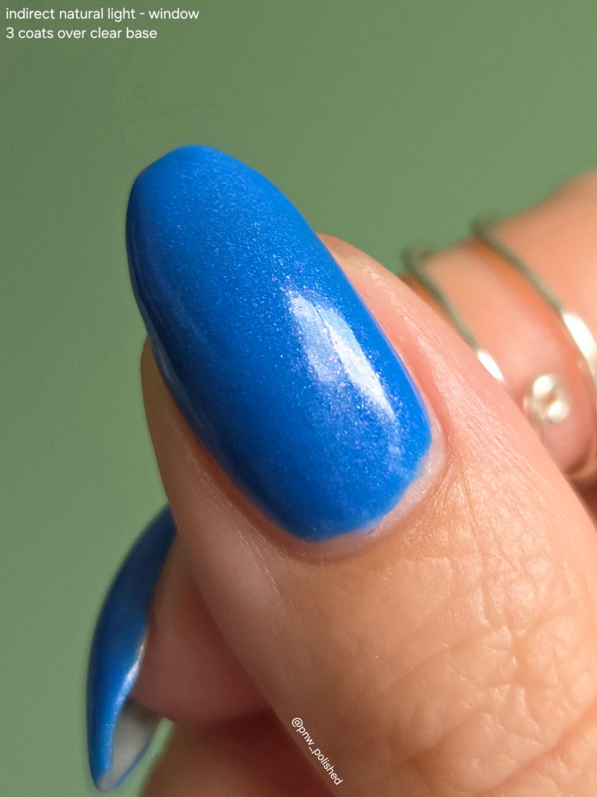 A close-up of a fingernail painted in a vivid medium blue polish, with part of another blue-painted nail visible at the lower left. Thin gold rings are worn on the fingers against a soft green background.