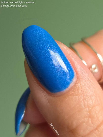 A close-up of a fingernail painted in a vivid medium blue polish, with part of another blue-painted nail visible at the lower left. Thin gold rings are worn on the fingers against a soft green background.