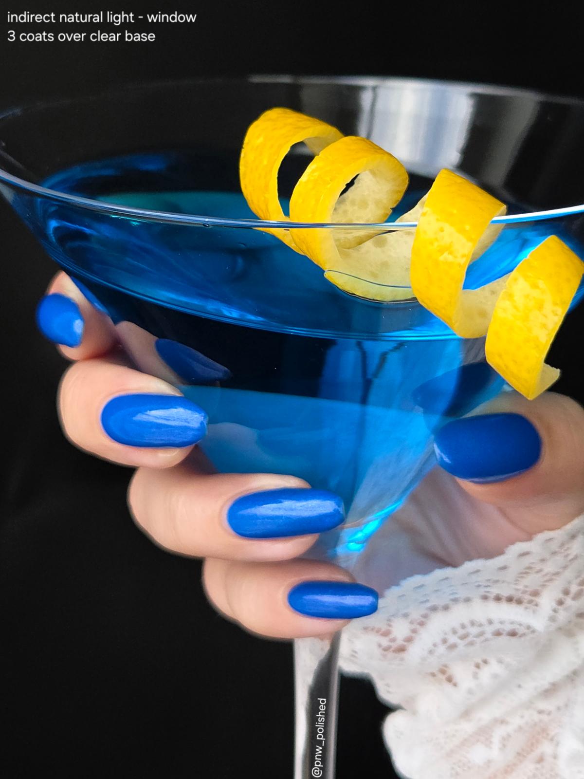 A close-up of a hand with bright royal blue nail polish holding a martini glass filled with vivid blue liquid and garnished with a curled yellow lemon peel. The background is black, and small text in the top left reads 'indirect natural light - window 3 coats over clear base'.
