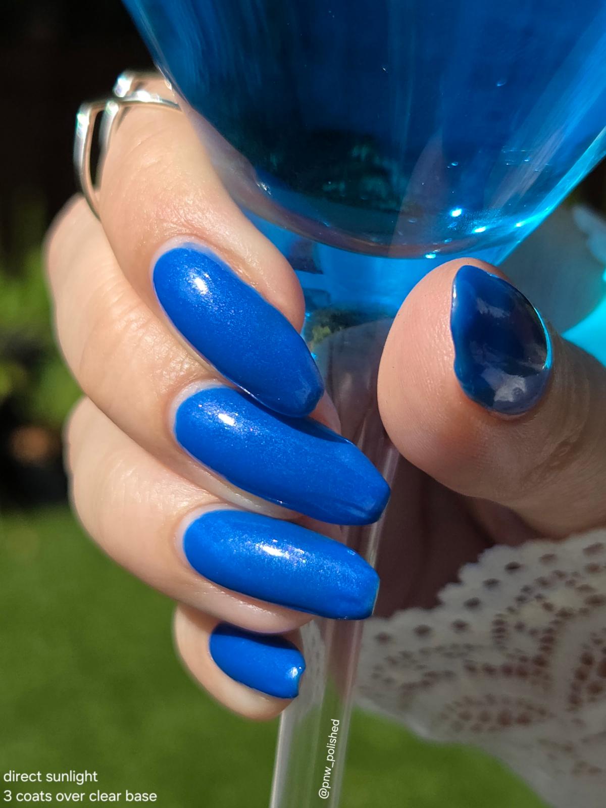 A close-up of a hand with long, glossy cobalt blue manicured nails holding a clear glass filled with bright blue liquid. The background is a soft-focus outdoor green lawn, and the text overlay at the bottom left reads 'direct sunlight 3 coats over clear base'.