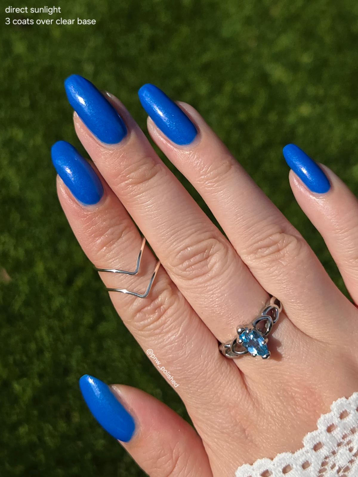 A close-up of a hand with long, glossy cobalt blue manicured nails, wearing a silver ring with a blue gemstone and a thin silver chevron ring, set against a blurred green grass background. Text in the top-left corner reads 'direct sunlight 3 coats over clear base'.